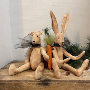 Peut inclure: Deux jouets en peluche de style primitif faits à la main, un ours et un lapin, assis sur une caisse en bois. L'ours porte un ruban noir et un nœud en tulle noir. Le lapin tient une carotte. La caisse porte les mots "Welcome-Cozy" gravés sur le devant.