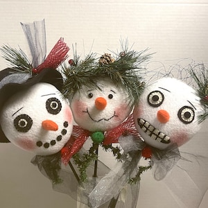 Könnte beinhalten: Drei Schneemann-Ornamente mit unterschiedlichen Gesichtsausdrücken. Der Schneemann links trägt einen schwarzen Hut und einen rot-weißen Schal. Der Schneemann in der Mitte hat einen grün-roten Schal. Der Schneemann rechts hat einen schwarz-weißen Schal.
