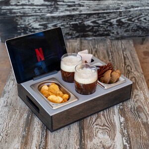 May include: A wooden serving tray with compartments for snacks and drinks. The tray has a built-in tablet holder and two glasses of beer. The tray also has a compartment for chips and a compartment for snacks. The tray is on a wooden table.
