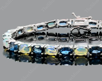 May include: A silver bracelet with alternating oval gemstones in blue and opalescent white. The bracelet has a secure clasp and is displayed on a reflective surface.