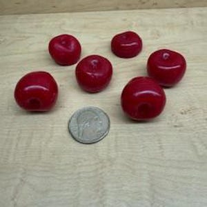 May include: Six small, glossy red apple-shaped beads are arranged on a light wood surface, with a U.S. quarter for size comparison. The beads are a vibrant shade of red, and each has a small indentation at the top.