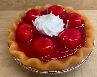May include: A red cherry pie candle with a white whipped cream topping. The pie is in a silver foil pan with a fluted crust.
