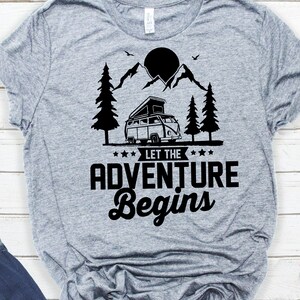 May include: Grey t-shirt with a black graphic of a camper van driving through a mountain landscape with a sun and trees. The text "Let the Adventure Begin" is below the camper van.