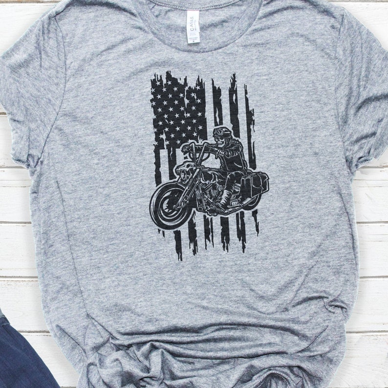 May include: A gray t-shirt with a black and white graphic of a motorcycle rider with a skull helmet riding in front of a distressed American flag.