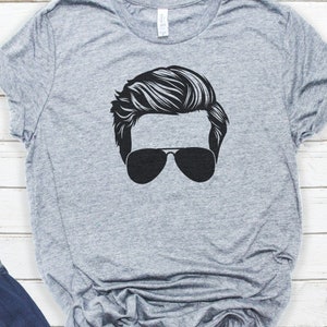 May include: A grey t-shirt featuring a black silhouette of a person wearing sunglasses. The person has a hairstyle with a side parting.