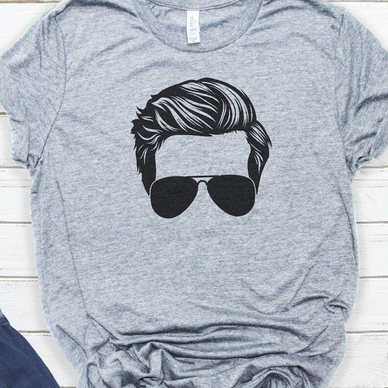 May include: A grey t-shirt featuring a black silhouette of a person wearing sunglasses. The person has a hairstyle with a side parting.