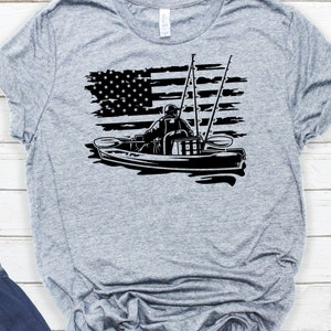 May include: A gray t-shirt with a black silhouette of a person kayaking with fishing rods. The kayak is in front of a distressed American flag.
