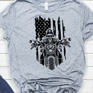May include: A grey t-shirt featuring a black and white graphic of a skeleton riding a motorbike in front of a distressed American flag.