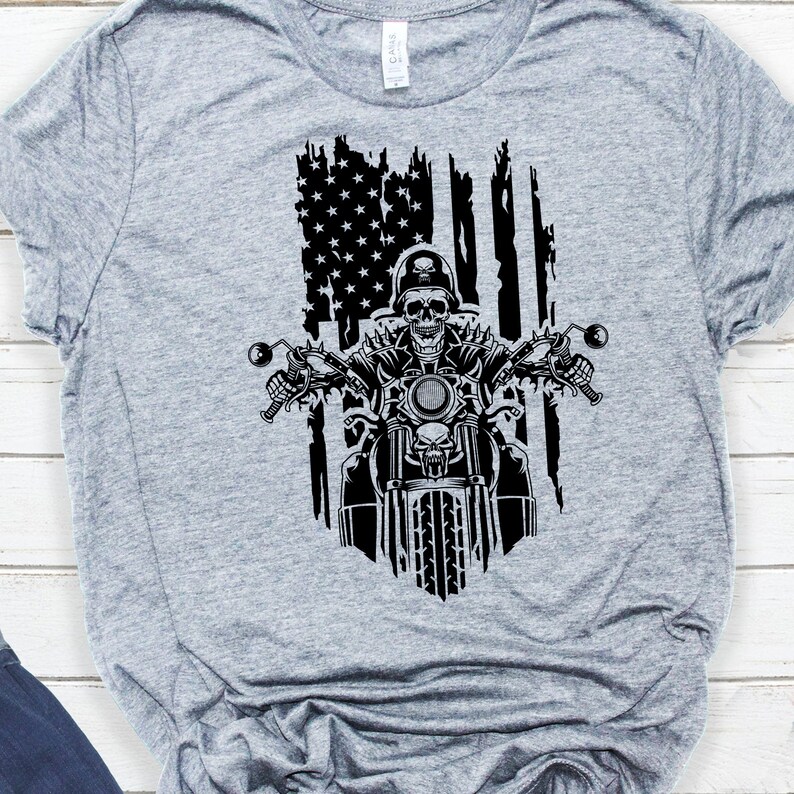 May include: A grey t-shirt featuring a black and white graphic of a skeleton riding a motorbike in front of a distressed American flag.