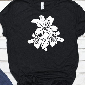 Puede incluir: Camiseta negra con un dise&ntilde;o floral blanco. El gr&aacute;fico es una ilustraci&oacute;n estilizada de varios lirios agrupados. La camiseta est&aacute; hecha de un material suave y tiene un cuello redondo cl&aacute;sico.