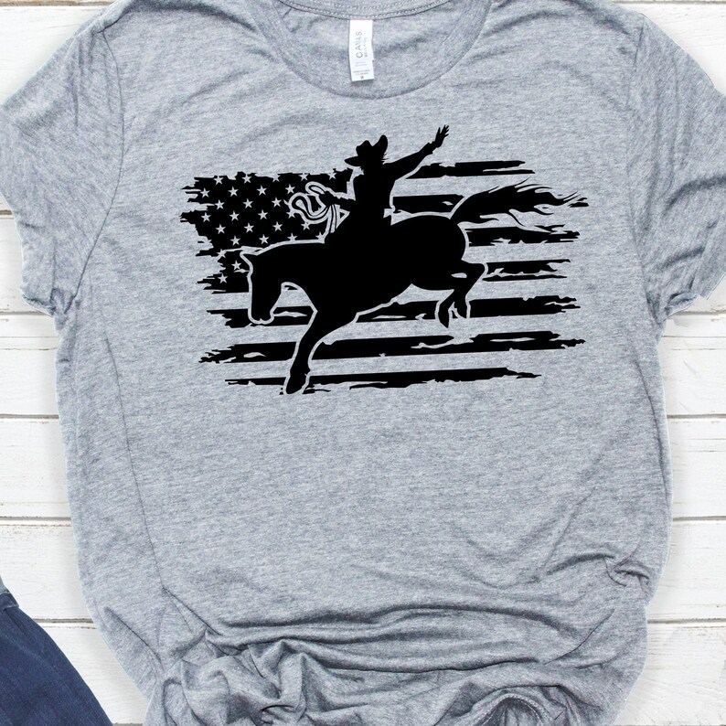 May include: A gray t-shirt with a black silhouette of a cowboy riding a bucking horse in front of a distressed American flag.