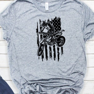 K&ouml;nnte beinhalten: Ein graues T-Shirt mit einer schwarz-wei&szlig;en Grafik eines Motocross-Fahrers, der &uuml;ber eine zerfetzte amerikanische Flagge springt.