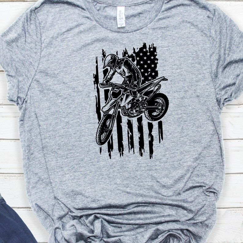 K&ouml;nnte beinhalten: Ein graues T-Shirt mit einer schwarz-wei&szlig;en Grafik eines Motocross-Fahrers, der &uuml;ber eine zerfetzte amerikanische Flagge springt.