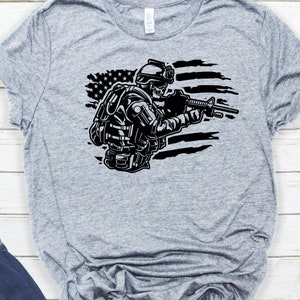 May include: A gray t-shirt with a black and white graphic of a soldier holding a rifle in front of a distressed American flag.