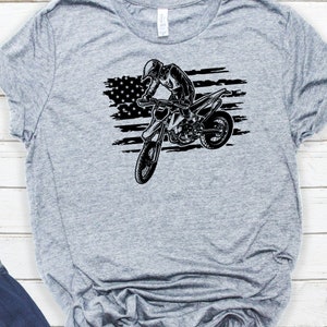 K&ouml;nnte beinhalten: Ein graues T-Shirt mit einer schwarz-wei&szlig;en Grafik einer Person, die ein Dirtbike vor einer amerikanischen Flagge f&auml;hrt.