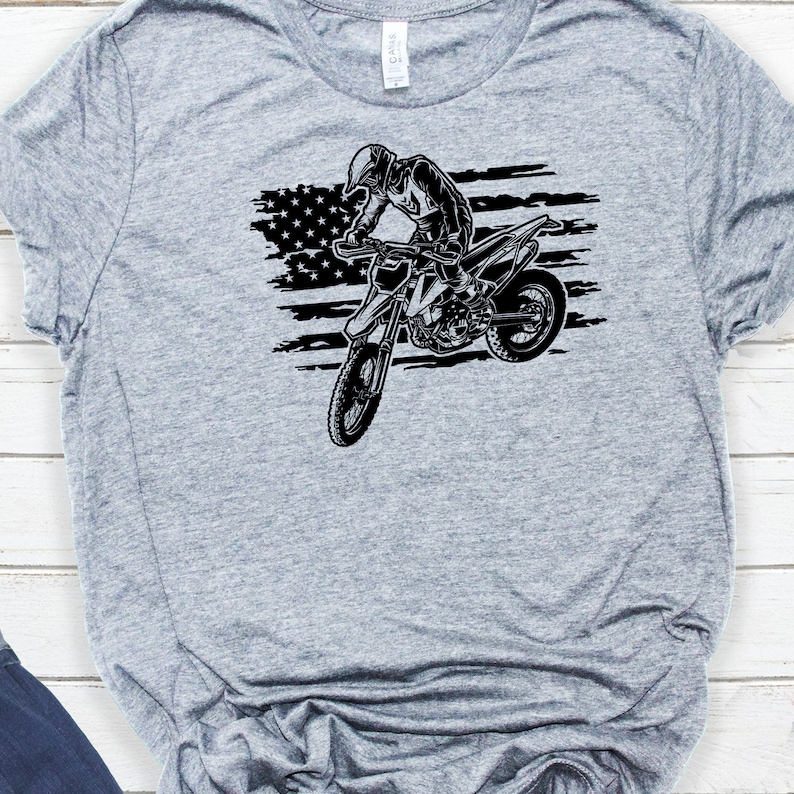 K&ouml;nnte beinhalten: Ein graues T-Shirt mit einer schwarz-wei&szlig;en Grafik einer Person, die ein Dirtbike vor einer amerikanischen Flagge f&auml;hrt.