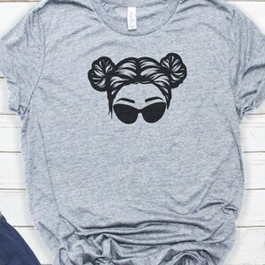 May include: A grey t-shirt with a black graphic of a woman with her hair in two buns and wearing sunglasses.