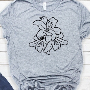 Puede incluir: Una camiseta gris con un dise&ntilde;o floral negro de lirios. El gr&aacute;fico es un contorno detallado de las flores, mostrando sus p&eacute;talos y estambres. La camiseta est&aacute; hecha de un material suave.