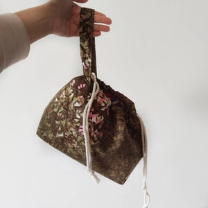 May include: A brown drawstring bag with a geometric pattern in shades of pink and green. The bag has a matching handle and a white drawstring closure. The bag is held up by a hand.