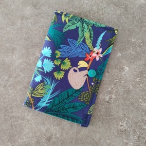 Puede incluir: Una cartera de tela con un estampado de jungla tropical en tonos azules, verdes y rosas. El diseño presenta un perezoso colgado de una rama. La cartera se asegura con un botón turquesa.