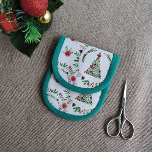 May include: A pair of white and green Christmas-themed needlework protectors with a floral and tree pattern. The protectors have a green border and are next to a pair of small scissors.