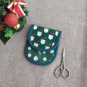May include: A green and white Christmas-themed pouch with a Santa Claus pattern and the word "HO" printed on it. A pair of small, silver scissors is next to the pouch. The pouch has a dark green trim.