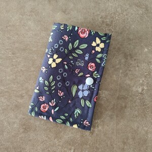 Könnte beinhalten: Ein marineblaues Portemonnaie mit floralem Muster und Knopfverschluss. Der Stoff zeigt ein Muster aus rosa, gelben und blauen Blumen mit grünen Blättern. Das Portemonnaie wird mit einem weißen Knopf verschlossen.