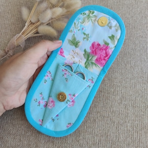 Puede incluir: Un estuche de tijeras hecho a mano con un estampado floral. El estuche es azul claro con un borde turquesa y un cierre magnético. El interior presenta un estampado floral y contiene un par de tijeras pequeñas.