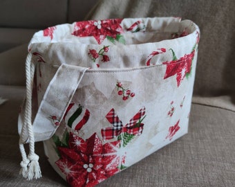 Puede incluir: Una cesta de almacenamiento rectangular de tela con cierre de cordón. La cesta presenta un fondo color crema con un patrón repetido de flores de pascua rojas, bastones de caramelo y lazos a cuadros. Se ve un cordón de cuerda blanca y un asa de tela.