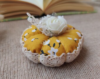 May include: A handmade pincushion shaped like a pumpkin, featuring a yellow floral fabric with white flowers. It has a cream lace trim and is topped with a white rose and lace embellishments. The pincushion is on a neutral-toned surface.