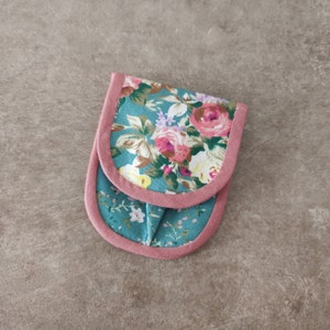 May include: A small pouch with a floral pattern on a teal background and pink trim. The pouch has a repeating pattern of pink and yellow roses and other flowers. The edges are outlined with a soft pink fabric.