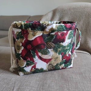 Può includere: Una borsa in tessuto con coulisse con un motivo natalizio festivo. La borsa presenta uno sfondo bianco con fiocchi rossi, agrifoglio verde e accenti dorati. Ha un cordoncino bianco e una piccola maniglia laterale.