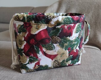 Puede incluir: Una bolsa de tela con cordón y un estampado navideño festivo. La bolsa presenta un fondo blanco con lazos rojos, acebo verde y detalles dorados. Tiene un cordón blanco y un asa pequeña en el lateral.