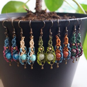 May include: A set of eight handmade macrame earrings with different colored gemstone beads. The earrings are made with a variety of colors, including blue, green, red, and brown. The earrings are hanging from a black pot.
