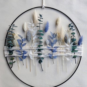 Op de afbeelding: Een zwarte metalen hoepel met een geweven macramé-ontwerp met gedroogde bloemen en groen in tinten blauw en wit.