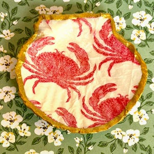 May include: A seashell-shaped decorative dish with a gold rim. The interior features a cream-colored background with a repeating pattern of red crabs. The dish is set against a green background with white floral accents.