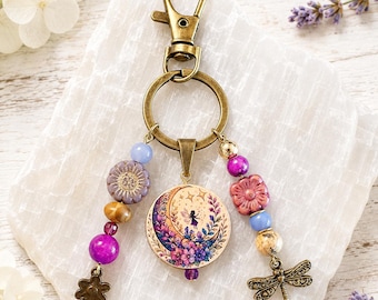 May include: A bronze-toned keychain featuring a central circular charm with a fairy and crescent moon illustration. Two beaded strands hang from the ring, decorated with purple, blue, and tan beads, a small bronze flower charm, and a dragonfly charm.
