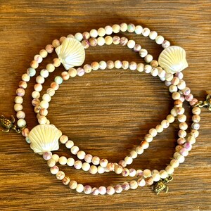 May include: A multi-strand necklace with cream-coloured beads, seashell and turtle charms. The beads have a speckled pattern with purple, orange, and blue hues. The seashells are pale yellow. The necklace is displayed on a wooden surface.