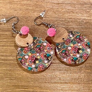 May include: A pair of dangle earrings with a wood and clear acrylic design. The earrings feature a pink bead and a floral pattern with butterflies and mushrooms.