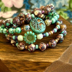 May include: Stacked beaded bracelets in shades of teal, purple, and brown. The bracelets feature various bead sizes and textures, including round and floral-shaped beads. A wooden block serves as a display.