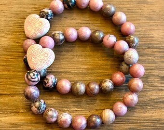 May include: Two pink and brown gemstone bracelets with white heart-shaped charms. The charms have a floral design.