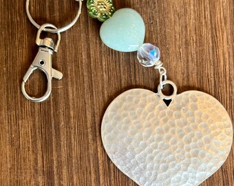 May include: A silver heart-shaped keychain with a hammered texture. The keychain has a silver ring and a silver lobster clasp. The keychain is decorated with green and gold beads and a clear crystal bead.