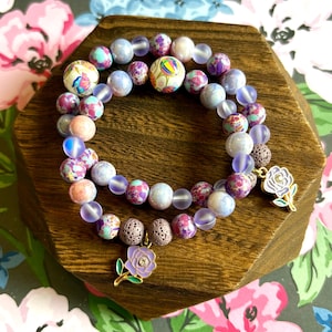 May include: Two beaded bracelets with a floral theme. The bracelets feature a mix of colorful beads, including patterned, iridescent, and round beads. Each bracelet has a gold-colored charm with a purple rose design. The bracelets are displayed on a wooden surface.