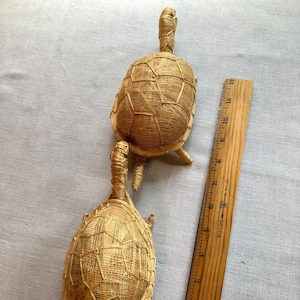 May include: Two woven straw turtle figurines on a white fabric background. The turtles are approximately 6 inches long and 4 inches wide. A wooden ruler is included for scale.