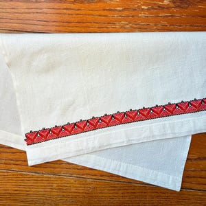 May include: White linen towel with a decorative border featuring a repeating pattern of red and black geometric shapes. The towel is folded, showcasing the embroidered design along one edge. The background is a wooden surface.