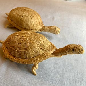 May include: Two woven straw turtle figurines. The turtles are brown and have a detailed shell pattern.