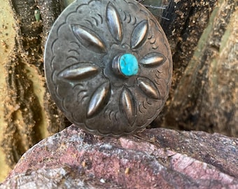 May include: A circular, silver-coloured metal button with an embossed floral pattern. A small, round turquoise stone is set in the centre. The button rests on a reddish-brown rock, with a cactus visible in the background.