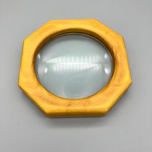 May include: A wooden magnifying glass with an octagonal frame. The frame is painted gold and surrounds a clear, convex lens. The magnifying glass is designed for reading or detailed inspection.