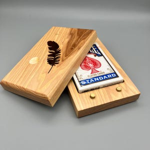 May include: A wooden playing card box with a feather design on the lid. The box is open, revealing a deck of playing cards. The box is made of light-colored wood and has brass-colored hardware.