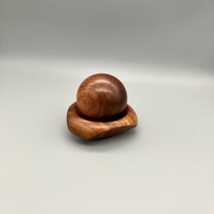 May include: A wooden sculpture comprising a sphere atop a uniquely shaped base. The wood is a warm brown with visible grain. The base has an organic, flowing form. The sculpture is likely handmade.